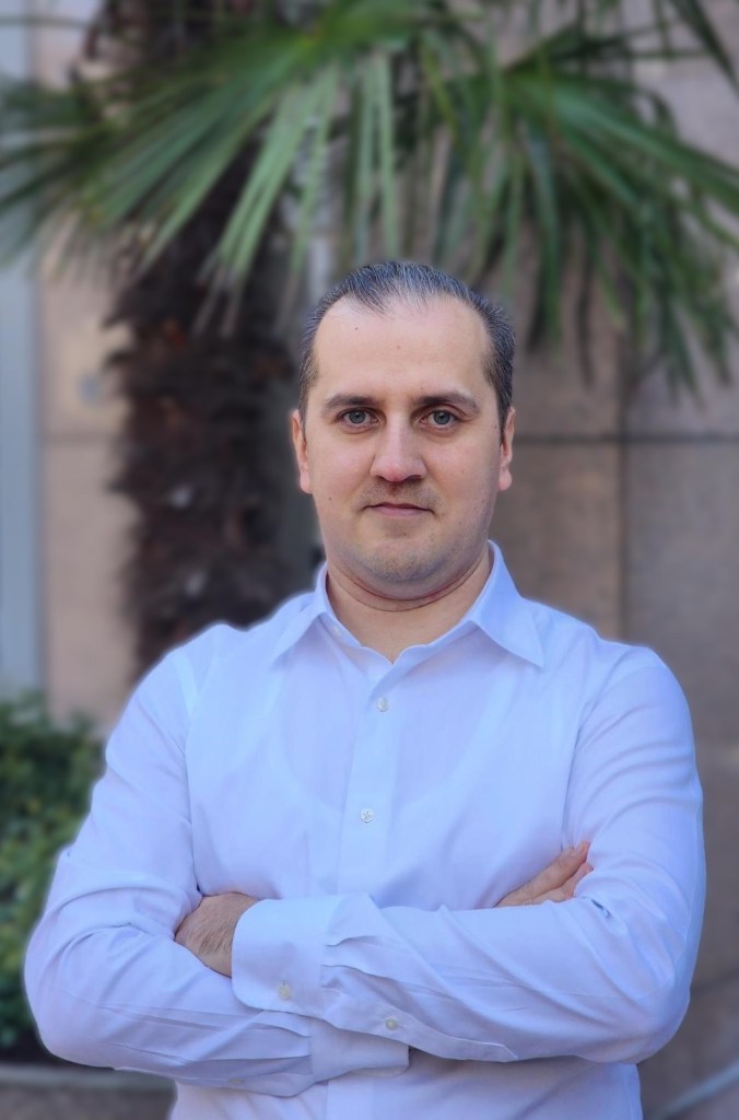 Photo of Fuat Ramazanov, a man wearing a long‑sleeved white dress shirt stands outdoors with arms crossed in front of a palm tree and a stone building.