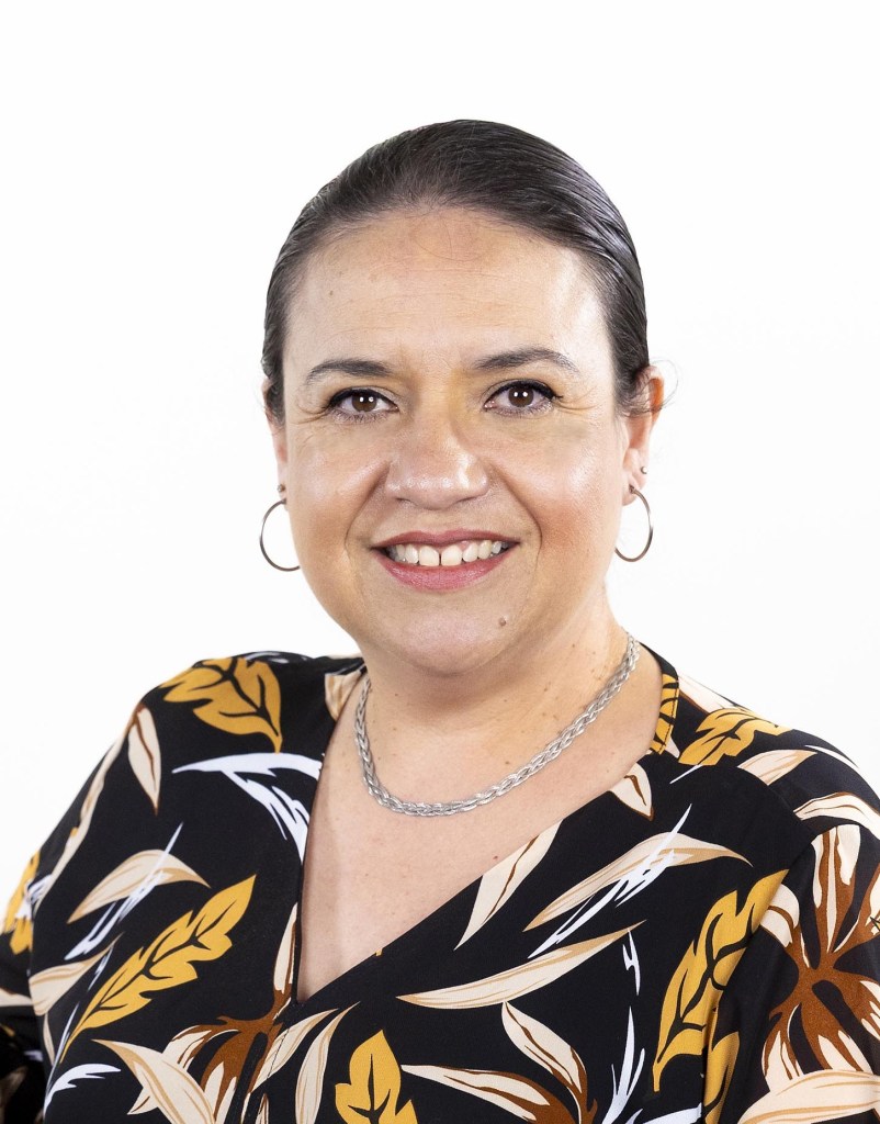 A head-and-shoulders photo of a woman with dark hair. She is wearing a dark top with a pattern in beige, yellow, and brown.