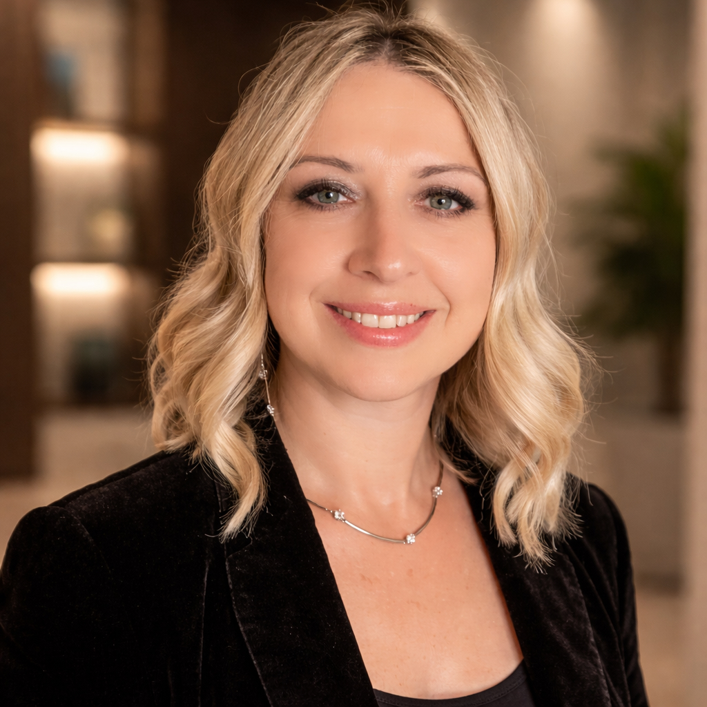 Close-up portrait of a person with shoulder-length, wavy light-colored hair, facing the camera. The person is wearing a dark blazer over a black top and a delicate necklace. The background is softly blurred, showing warm indoor lighting, shelving, and a plant, creating a professional, studio-like setting.